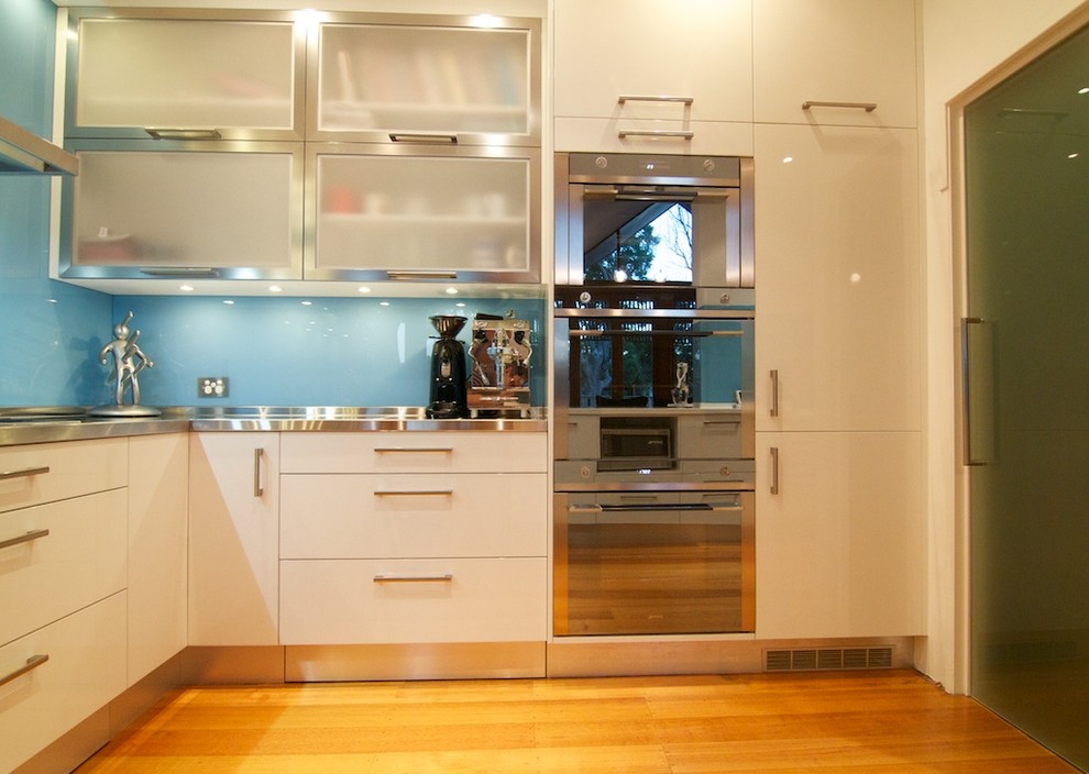 Inredning av ett modernt avskilt, mellanstort vit vitt l-kök, med en integrerad diskho, släta luckor, vita skåp, bänkskiva i rostfritt stål, blått stänkskydd, glaspanel som stänkskydd, rostfria vitvaror, ljust trägolv och en köksö