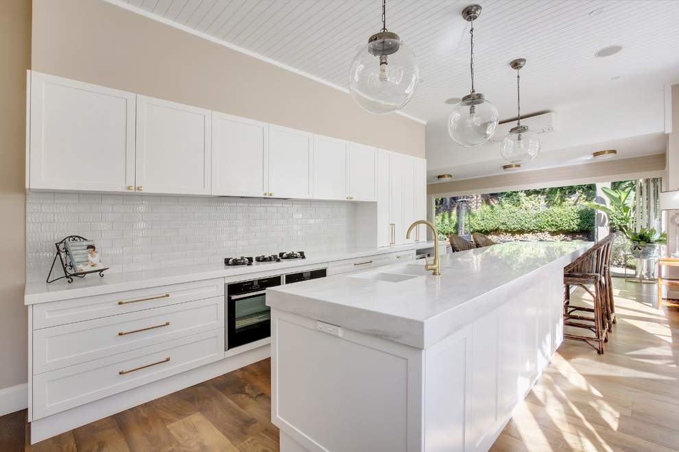 Expansive l-shaped open plan kitchen in Sydney with a submerged sink, shaker cabinets, white cabinets, composite countertops, white splashback, metro tiled splashback, stainless steel appliances, medium hardwood flooring, an island, brown floors and white worktops.