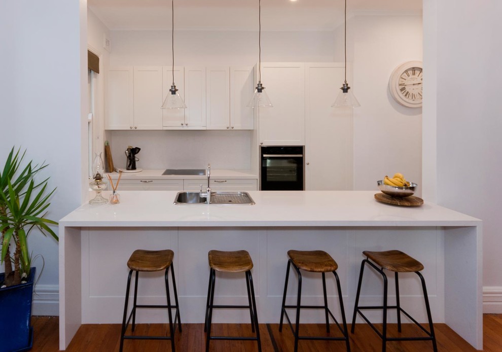 Zweizeilige, Kleine Moderne Küche mit Doppelwaschbecken, Schrankfronten im Shaker-Stil, weißen Schränken, Quarzwerkstein-Arbeitsplatte, Küchenrückwand in Weiß, Glasrückwand, braunem Holzboden und Halbinsel in Sydney