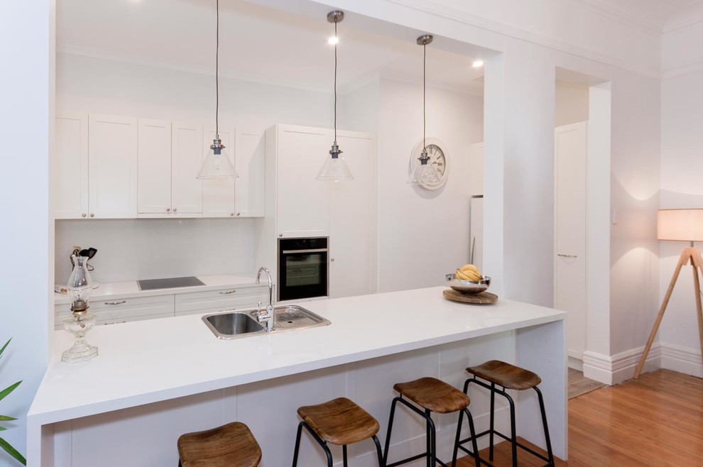 Photo of a small contemporary galley kitchen in Sydney with a double-bowl sink, shaker cabinets, white cabinets, engineered stone countertops, white splashback, glass sheet splashback, medium hardwood flooring and a breakfast bar.