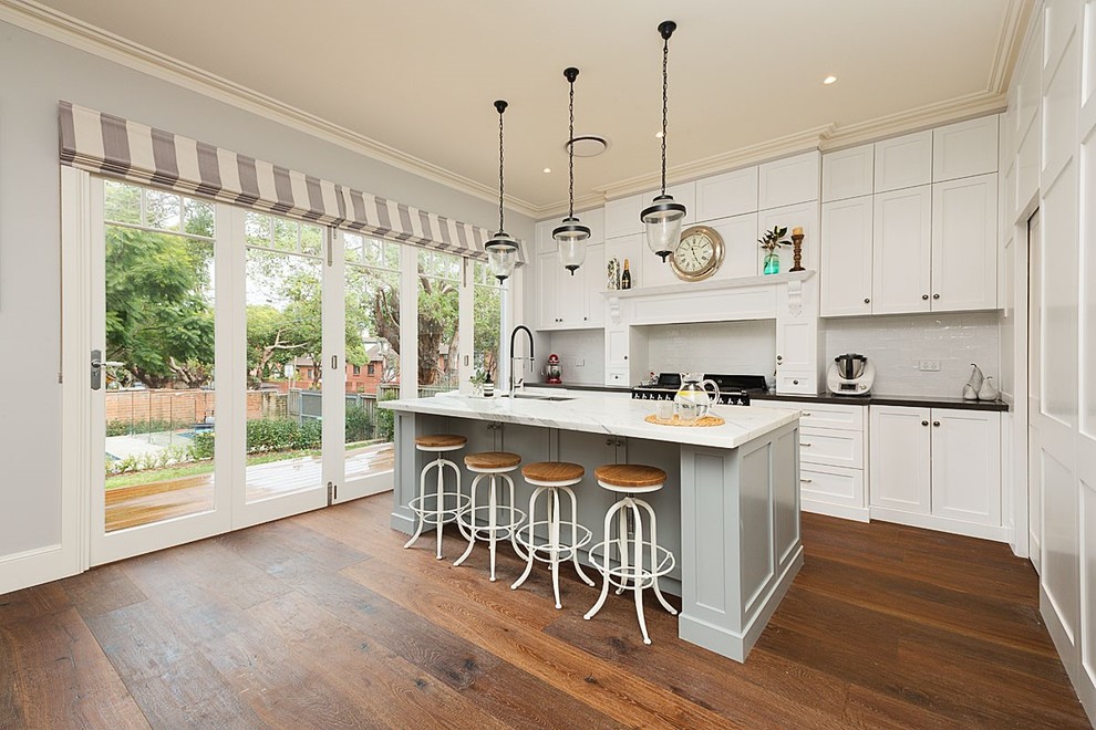 Eat-in kitchen - large traditional galley medium tone wood floor and brown floor eat-in kitchen idea in Sydney with a drop-in sink, shaker cabinets, white cabinets, marble countertops, white backsplash, ceramic backsplash, stainless steel appliances and an island