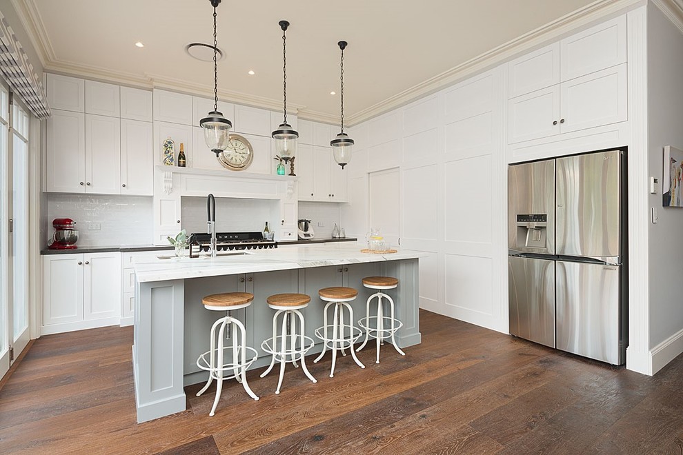 Photo of a large traditional galley kitchen/diner in Sydney with a built-in sink, shaker cabinets, white cabinets, marble worktops, white splashback, ceramic splashback, stainless steel appliances, medium hardwood flooring, an island and brown floors.