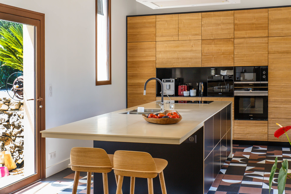 Photo of a contemporary kitchen in Sussex with a double-bowl sink, flat-panel cabinets, light wood cabinets, black splashback, black appliances, an island, multi-coloured floors and beige worktops.