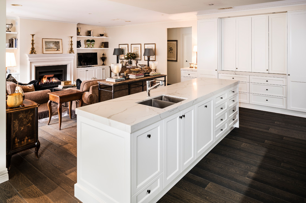 Inspiration for a large traditional galley open plan kitchen in Melbourne with a double-bowl sink, recessed-panel cabinets, brown cabinets, engineered stone countertops, white splashback, stone slab splashback, black appliances, dark hardwood flooring, an island, brown floors and white worktops.