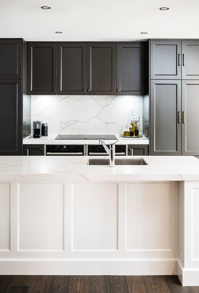 Klassisk inredning av ett stort vit vitt kök, med en dubbel diskho, luckor med infälld panel, bruna skåp, bänkskiva i kvarts, vitt stänkskydd, stänkskydd i sten, svarta vitvaror, mörkt trägolv, en köksö och brunt golv