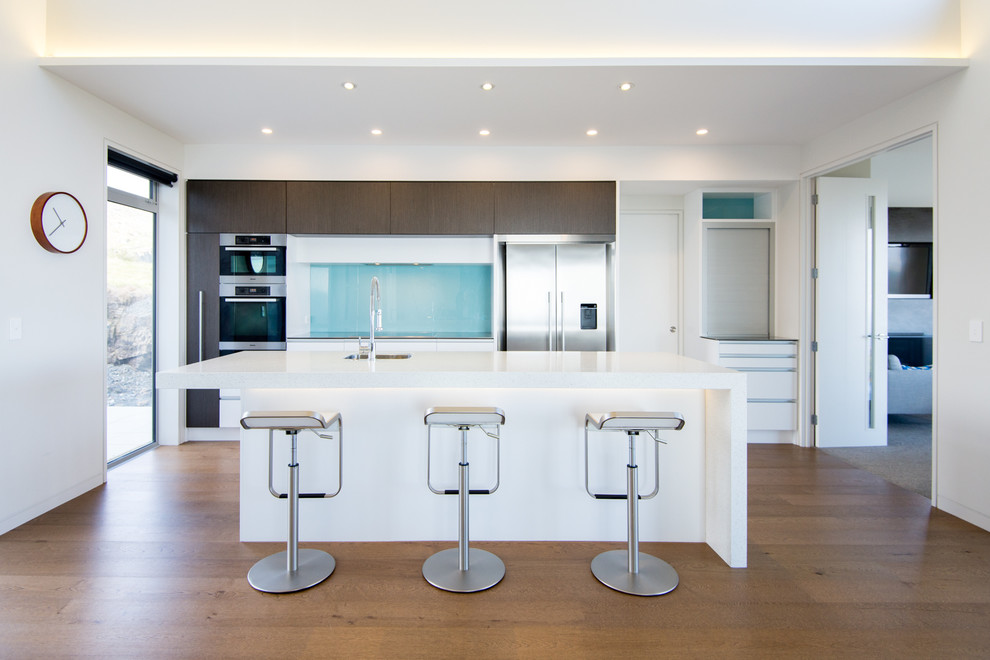 This is an example of a medium sized contemporary galley open plan kitchen in Auckland with a submerged sink, flat-panel cabinets, white cabinets, blue splashback, glass sheet splashback, stainless steel appliances, medium hardwood flooring and an island.