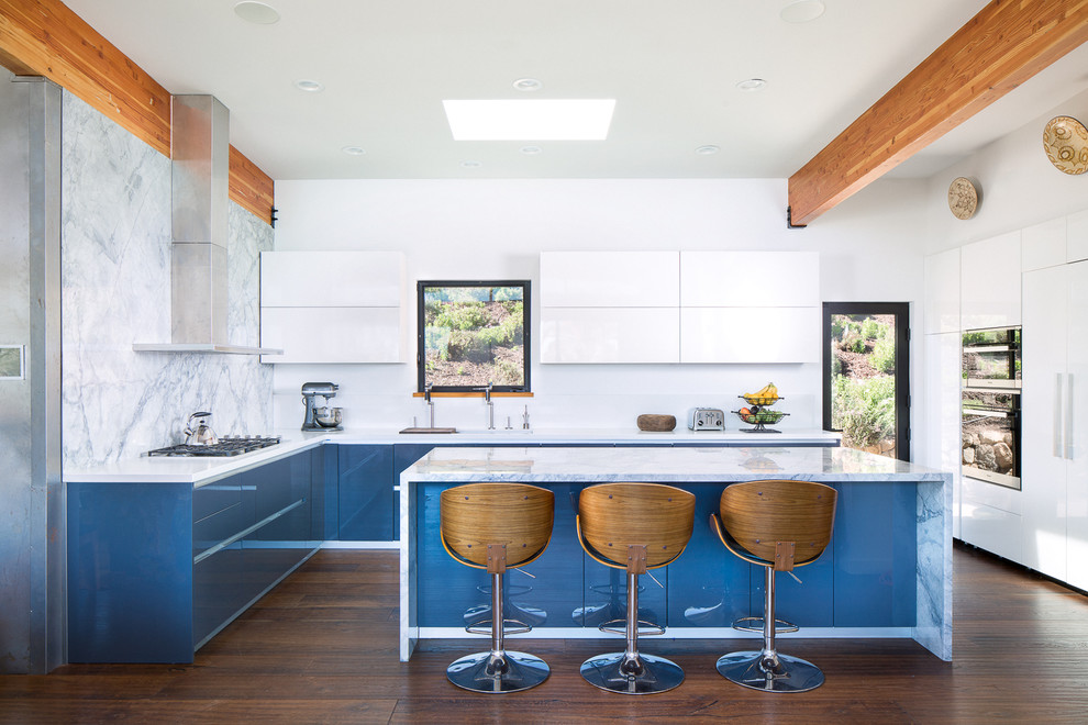 Inredning av ett modernt l-kök, med släta luckor, blå skåp, vitt stänkskydd, stänkskydd i marmor, mörkt trägolv, en köksö och brunt golv