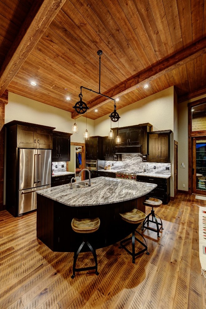 This is an example of a rustic l-shaped open plan kitchen in Denver with a double-bowl sink, recessed-panel cabinets, grey splashback, marble splashback, stainless steel appliances, medium hardwood flooring, an island and black cabinets.