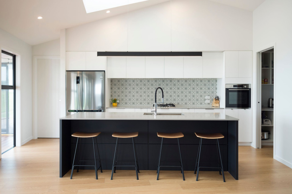 Contemporary galley kitchen in Auckland with flat-panel cabinets, white cabinets, grey splashback, mosaic tiled splashback, stainless steel appliances, light hardwood flooring, an island, beige floors, grey worktops and a vaulted ceiling.