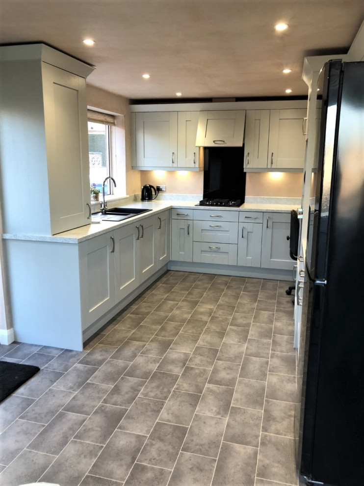 Example of a mid-sized classic u-shaped gray floor eat-in kitchen design in West Midlands with shaker cabinets, gray cabinets, laminate countertops, black backsplash, glass sheet backsplash, black appliances, no island and white countertops