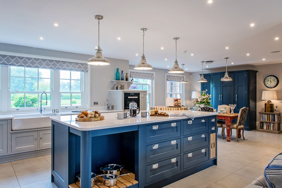 Design ideas for a large traditional open plan kitchen in Surrey with a belfast sink, shaker cabinets, blue cabinets, quartz worktops, white splashback, ceramic splashback, stainless steel appliances, porcelain flooring, an island and grey floors.