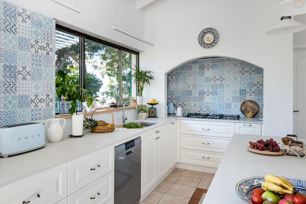 Foto på ett mellanstort medelhavsstil vit l-kök, med en undermonterad diskho, bänkskiva i koppar, blått stänkskydd, stänkskydd i keramik, rostfria vitvaror, klinkergolv i keramik, en köksö, luckor med upphöjd panel, vita skåp och beiget golv