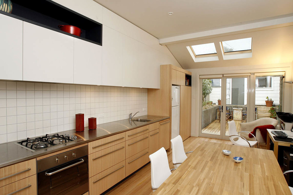 Foto på ett litet funkis linjärt kök och matrum, med en nedsänkt diskho, skåp i mellenmörkt trä, bänkskiva i rostfritt stål, vitt stänkskydd, stänkskydd i keramik, vita vitvaror, mellanmörkt trägolv och en köksö