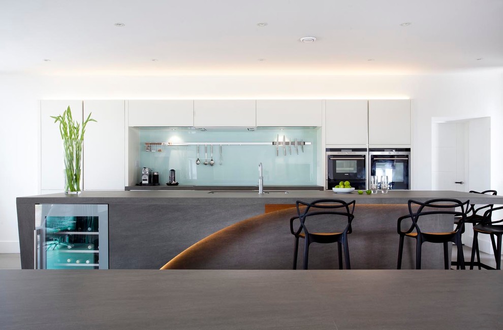Photo of an expansive contemporary u-shaped open plan kitchen in Other with an integrated sink, flat-panel cabinets, white cabinets, engineered stone countertops, green splashback, glass sheet splashback, stainless steel appliances, ceramic flooring and an island.