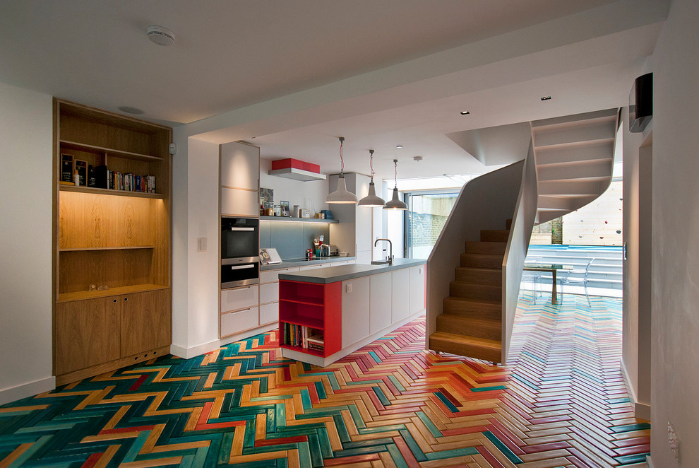 Свежая идея для дизайна: параллельная кухня-гостиная в стиле фьюжн с разноцветным полом, плоскими фасадами, белыми фасадами, столешницей из бетона, серым фартуком, черной техникой, деревянным полом и островом - отличное фото интерьера