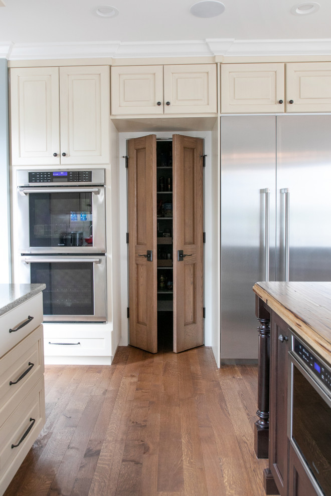 This is an example of a large classic grey and cream l-shaped kitchen pantry in Milwaukee with a belfast sink, raised-panel cabinets, stainless steel appliances, medium hardwood flooring, an island, beige cabinets, granite worktops, white splashback, metro tiled splashback, brown floors and grey worktops.