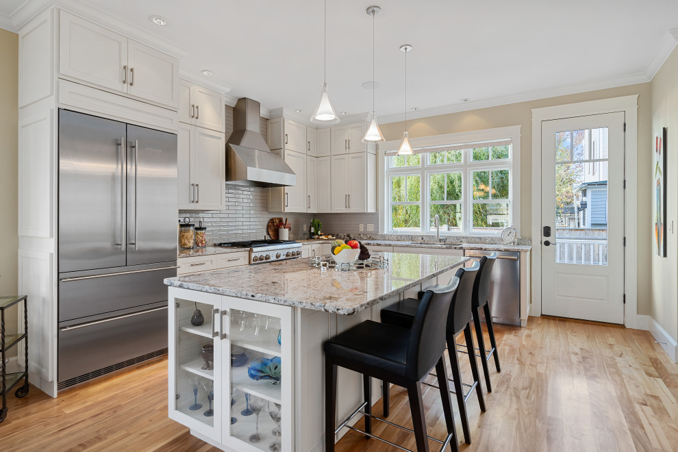 Large transitional l-shaped medium tone wood floor and brown floor kitchen photo in Portland Maine with an undermount sink, shaker cabinets, white cabinets, granite countertops, white backsplash, stainless steel appliances, an island and gray countertops