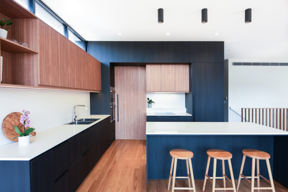 Inspiration for a large modern l-shaped open plan kitchen in Sydney with a built-in sink, flat-panel cabinets, black cabinets, engineered stone countertops, white splashback, stone slab splashback, black appliances, medium hardwood flooring, an island, brown floors and white worktops.