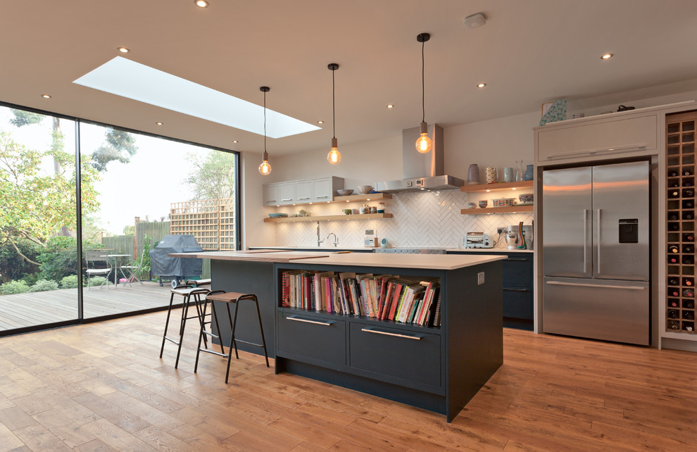 Mid-sized trendy single-wall medium tone wood floor and brown floor open concept kitchen photo in Essex with an undermount sink, flat-panel cabinets, blue cabinets, white backsplash, porcelain backsplash, stainless steel appliances and an island
