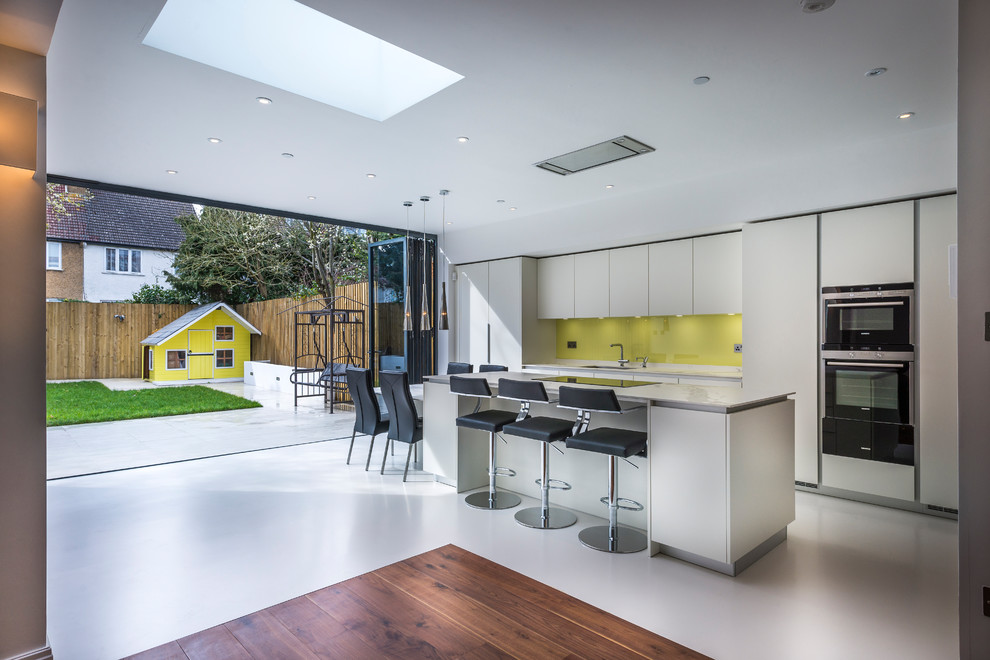 Идея дизайна: параллельная кухня-гостиная в современном стиле с плоскими фасадами, белыми фасадами, желтым фартуком, фартуком из стекла, островом, белым полом и белой столешницей