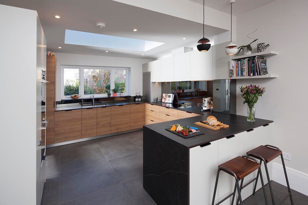 This is an example of a medium sized victorian u-shaped open plan kitchen in London with a built-in sink, flat-panel cabinets, medium wood cabinets, integrated appliances, ceramic flooring, a breakfast bar, grey floors and black worktops.