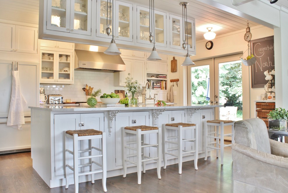 Foto på ett lantligt kök, med luckor med glaspanel, vita skåp, vitt stänkskydd, stänkskydd i tunnelbanekakel, integrerade vitvaror, mörkt trägolv och en köksö