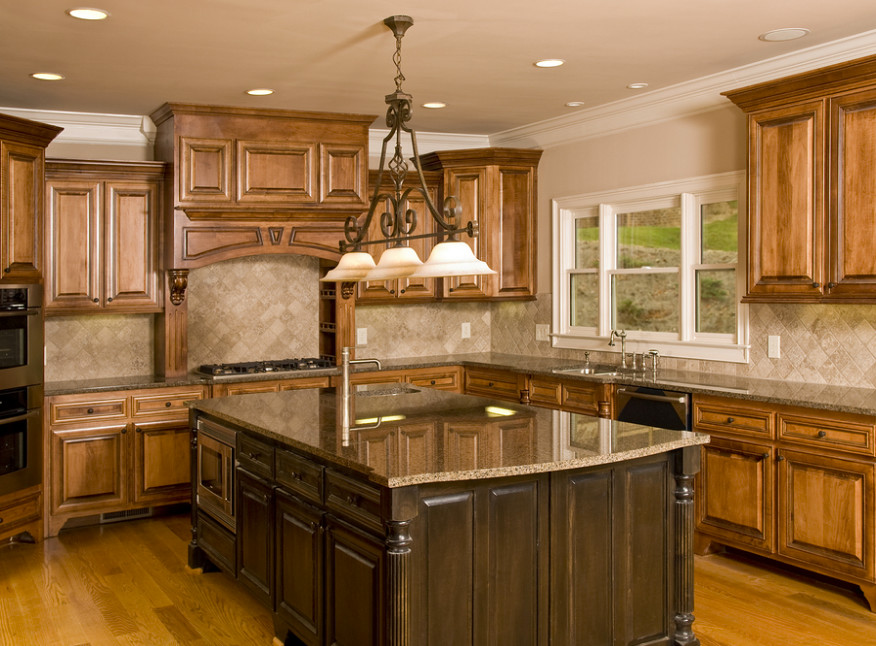 Inredning av ett klassiskt mellanstort brun brunt kök, med en dubbel diskho, luckor med upphöjd panel, skåp i ljust trä, granitbänkskiva, brunt stänkskydd, stänkskydd i stenkakel, rostfria vitvaror, mellanmörkt trägolv, en köksö och brunt golv