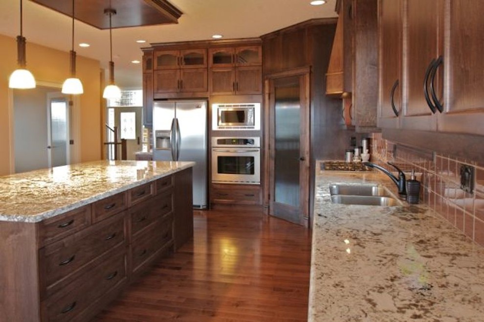 Photo of a medium sized traditional l-shaped kitchen in Calgary with a double-bowl sink, shaker cabinets, dark wood cabinets, granite worktops, brown splashback, ceramic splashback, stainless steel appliances, dark hardwood flooring, an island and brown floors.
