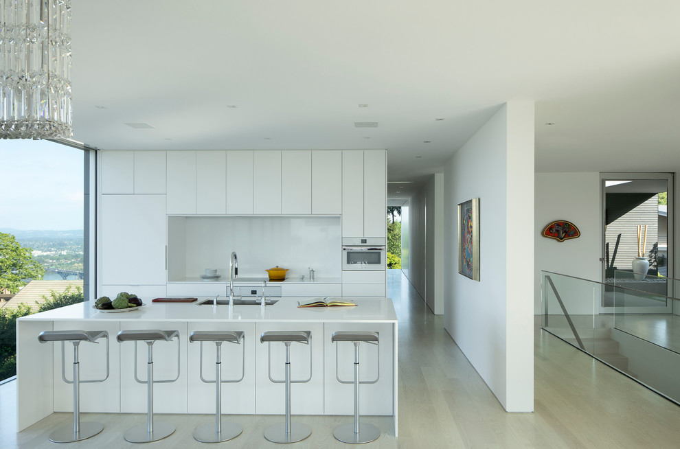 This is an example of a modern galley kitchen in Portland with flat-panel cabinets, white cabinets, integrated appliances, light hardwood flooring, an island, beige floors and white worktops.