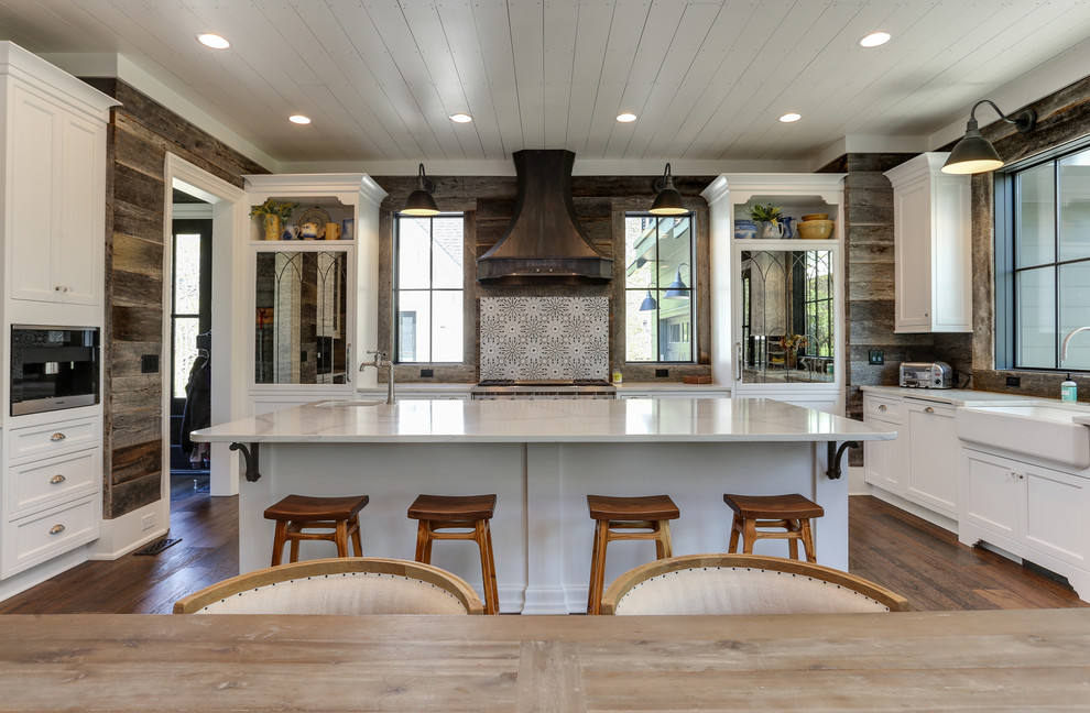 Bild på ett stort lantligt vit vitt kök, med en rustik diskho, luckor med profilerade fronter, vita skåp, bänkskiva i kvarts, brunt stänkskydd, integrerade vitvaror, mellanmörkt trägolv, en köksö och brunt golv