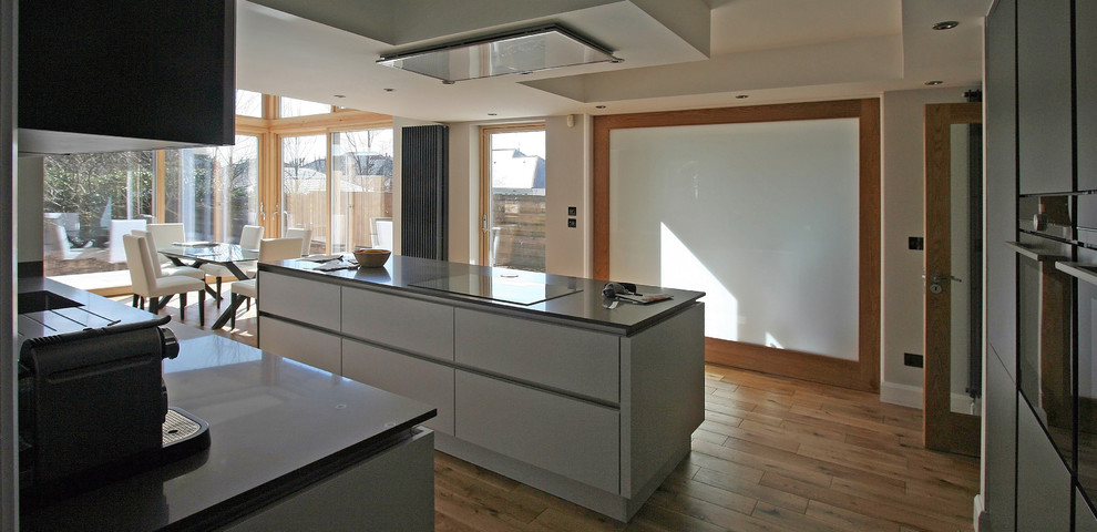This is an example of a medium sized modern galley open plan kitchen in Other with flat-panel cabinets, white cabinets, composite countertops, grey splashback, medium hardwood flooring, an island and grey worktops.
