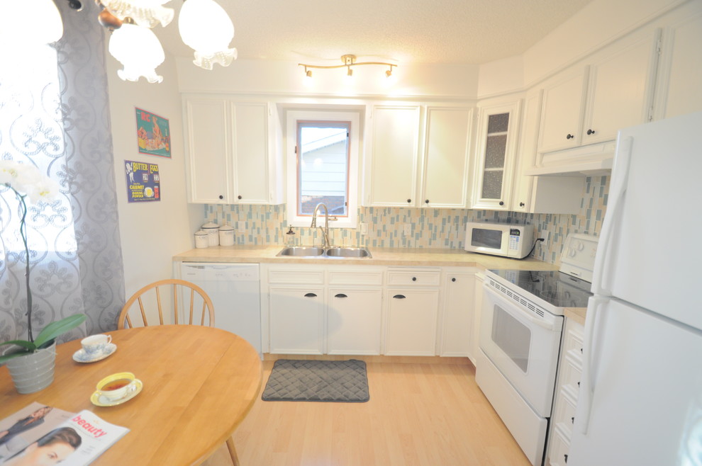 Kleine Shabby-Style Wohnküche in L-Form mit Einbauwaschbecken, Kassettenfronten, gelben Schränken, Laminat-Arbeitsplatte, Küchenrückwand in Blau, Rückwand aus Glasfliesen, weißen Elektrogeräten und hellem Holzboden in Calgary
