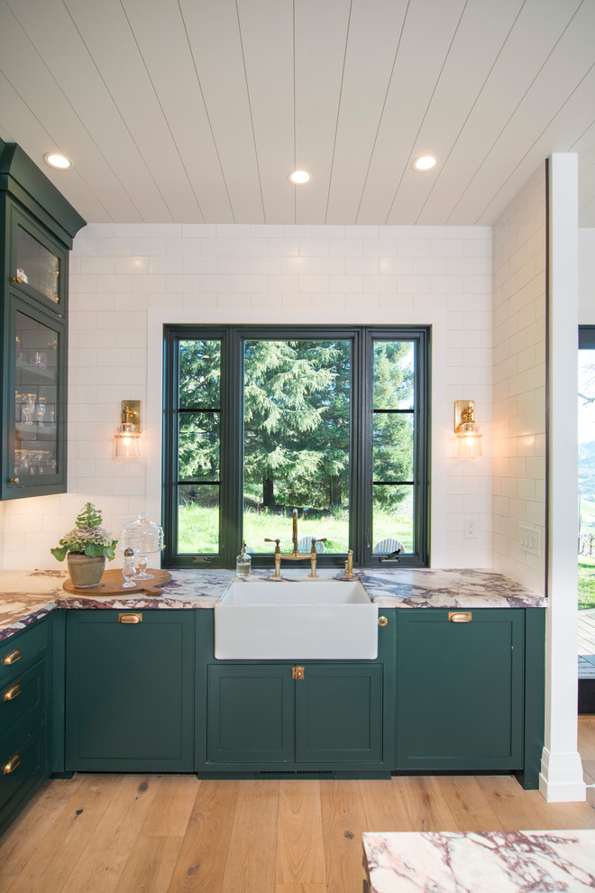 Ejemplo de cocina comedor de estilo de casa de campo grande con fregadero encastrado, armarios con paneles lisos, puertas de armario verdes, encimera de mármol, salpicadero blanco, salpicadero de azulejos de cerámica, electrodomésticos de acero inoxidable, suelo de madera en tonos medios, una isla, suelo marrón y encimeras blancas