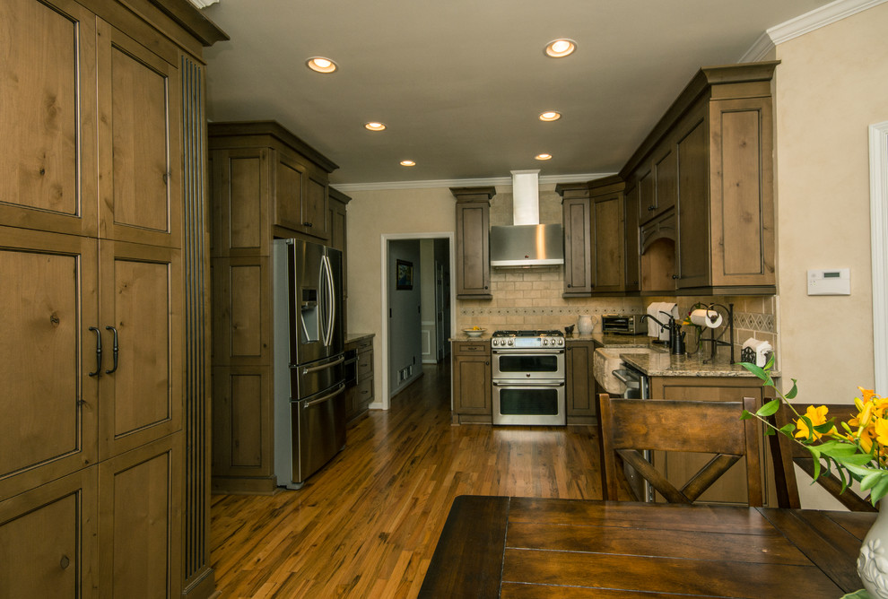 Example of a large classic l-shaped medium tone wood floor and brown floor enclosed kitchen design in Atlanta with a farmhouse sink, shaker cabinets, medium tone wood cabinets, granite countertops, beige backsplash, stone tile backsplash, stainless steel appliances and an island