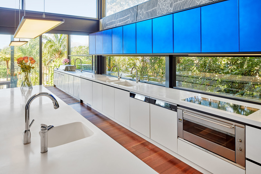 Offene, Große, Zweizeilige Moderne Küche mit flächenbündigen Schrankfronten, blauen Schränken, Mineralwerkstoff-Arbeitsplatte, Elektrogeräten mit Frontblende, braunem Holzboden, braunem Boden, integriertem Waschbecken, Rückwand-Fenster und Kücheninsel in Sydney