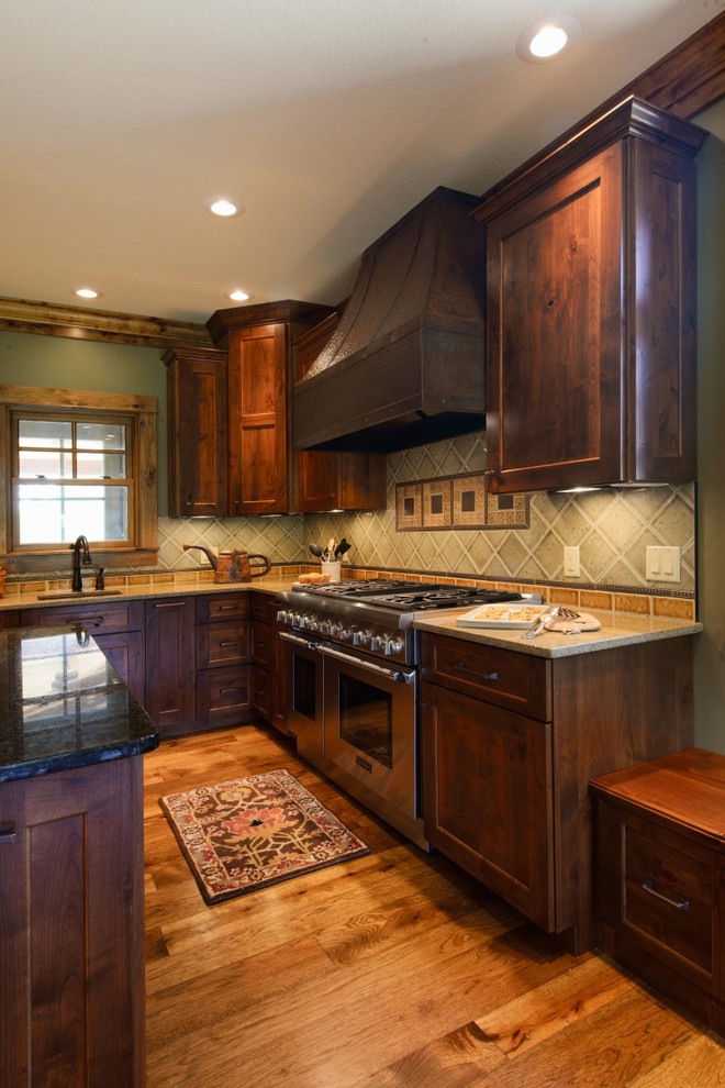 Lantlig inredning av ett kök, med en rustik diskho, luckor med infälld panel, skåp i slitet trä, granitbänkskiva, grönt stänkskydd, stänkskydd i porslinskakel, rostfria vitvaror, mellanmörkt trägolv och en köksö