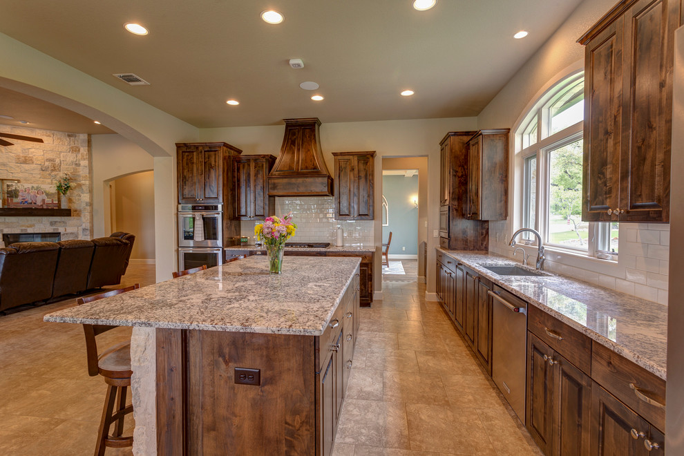 Example of a large mountain style l-shaped ceramic tile and beige floor open concept kitchen design in Austin with an undermount sink, raised-panel cabinets, dark wood cabinets, granite countertops, white backsplash, subway tile backsplash, stainless steel appliances, an island and multicolored countertops