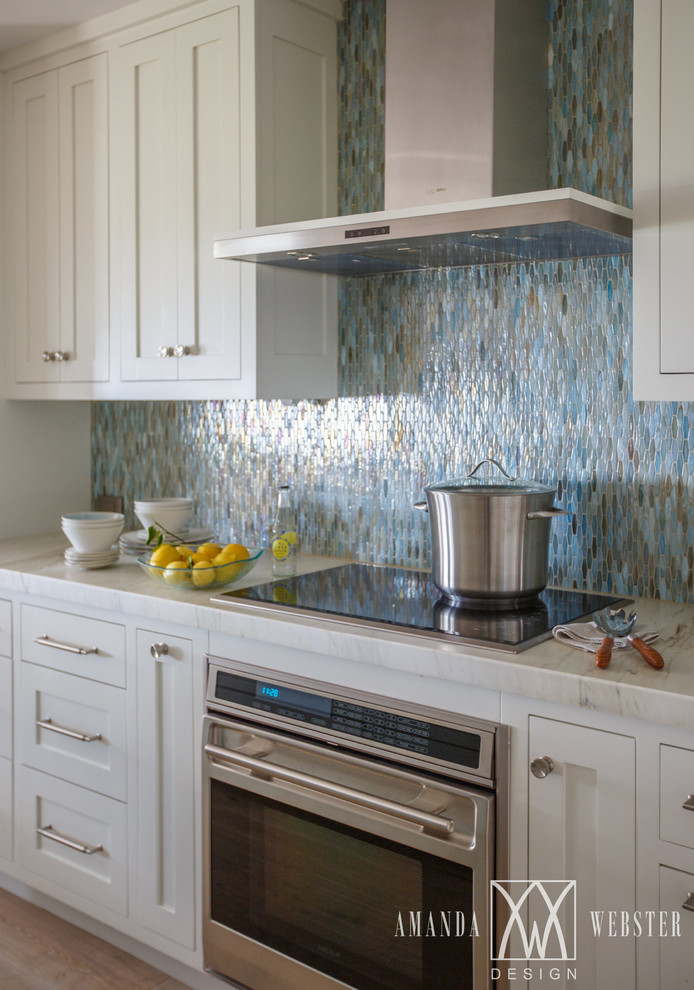 Maritim inredning av ett kök, med en undermonterad diskho, skåp i shakerstil, vita skåp, marmorbänkskiva, blått stänkskydd, stänkskydd i glaskakel, rostfria vitvaror, ljust trägolv och en köksö