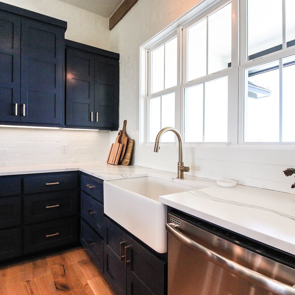 This is an example of a traditional l-shaped kitchen/diner in Dallas with a belfast sink, blue cabinets, engineered stone countertops, white splashback, brick splashback, stainless steel appliances, light hardwood flooring, an island and white worktops.