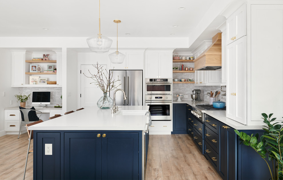 Design ideas for a large nautical l-shaped open plan kitchen in San Francisco with a belfast sink, shaker cabinets, white cabinets, composite countertops, grey splashback, stainless steel appliances, light hardwood flooring, an island, brown floors and white worktops.