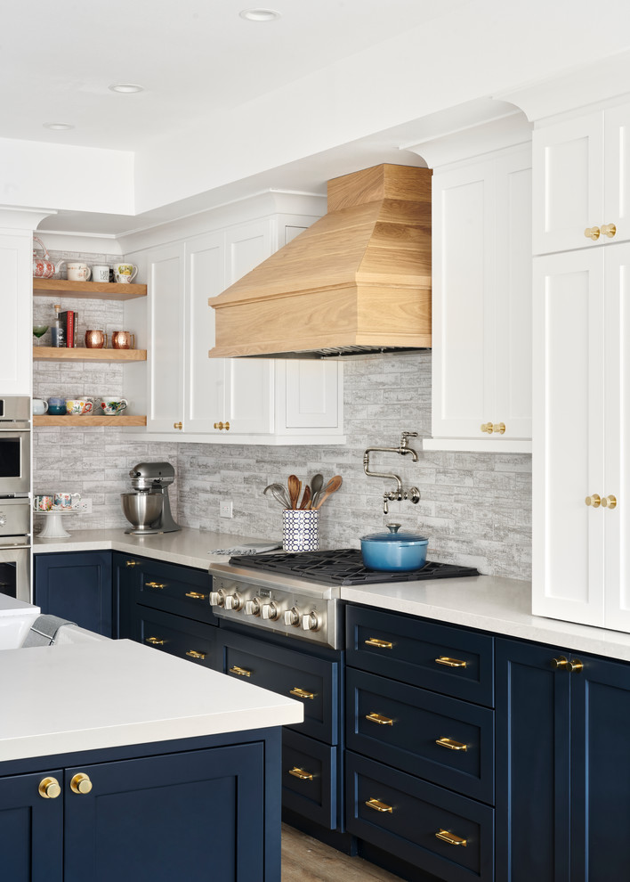 This is an example of a large nautical l-shaped open plan kitchen in San Francisco with a belfast sink, shaker cabinets, white cabinets, composite countertops, grey splashback, stainless steel appliances, light hardwood flooring, an island, brown floors and white worktops.