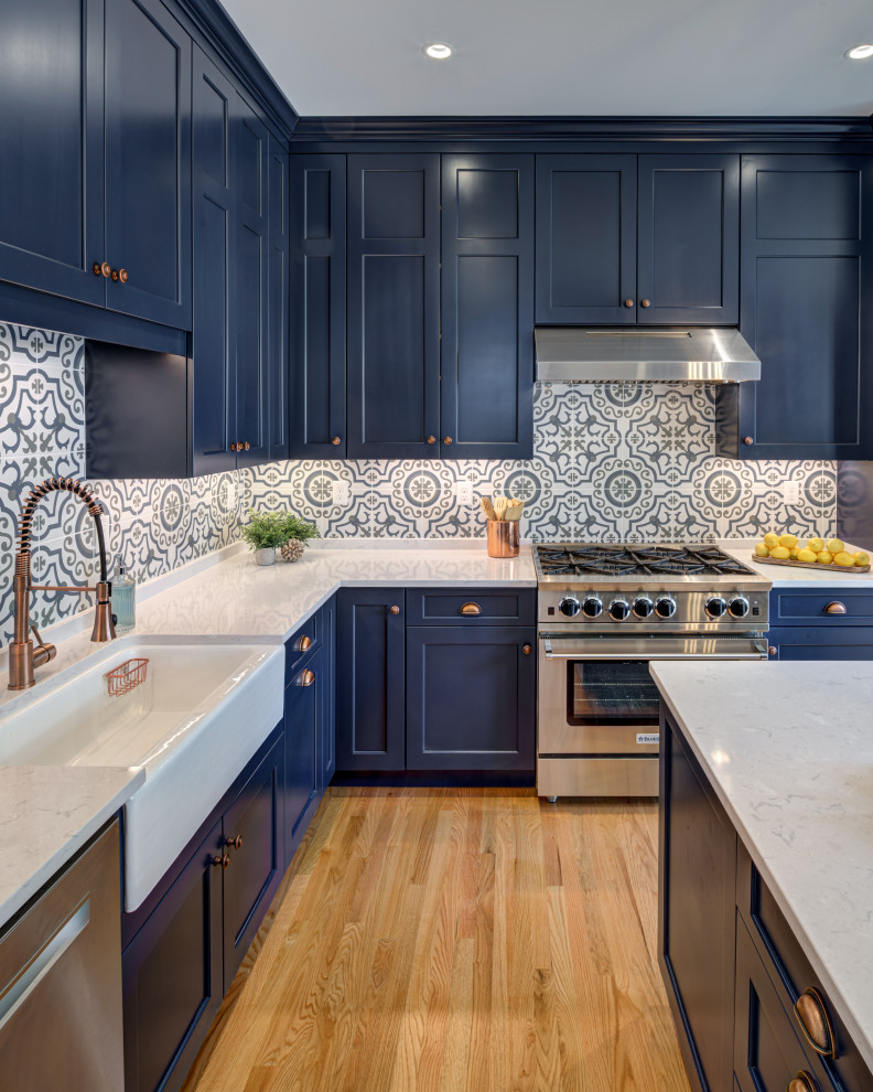Photo of a medium sized traditional l-shaped enclosed kitchen in DC Metro with a belfast sink, shaker cabinets, blue cabinets, engineered stone countertops, multi-coloured splashback, ceramic splashback, stainless steel appliances, medium hardwood flooring, an island, brown floors and white worktops.