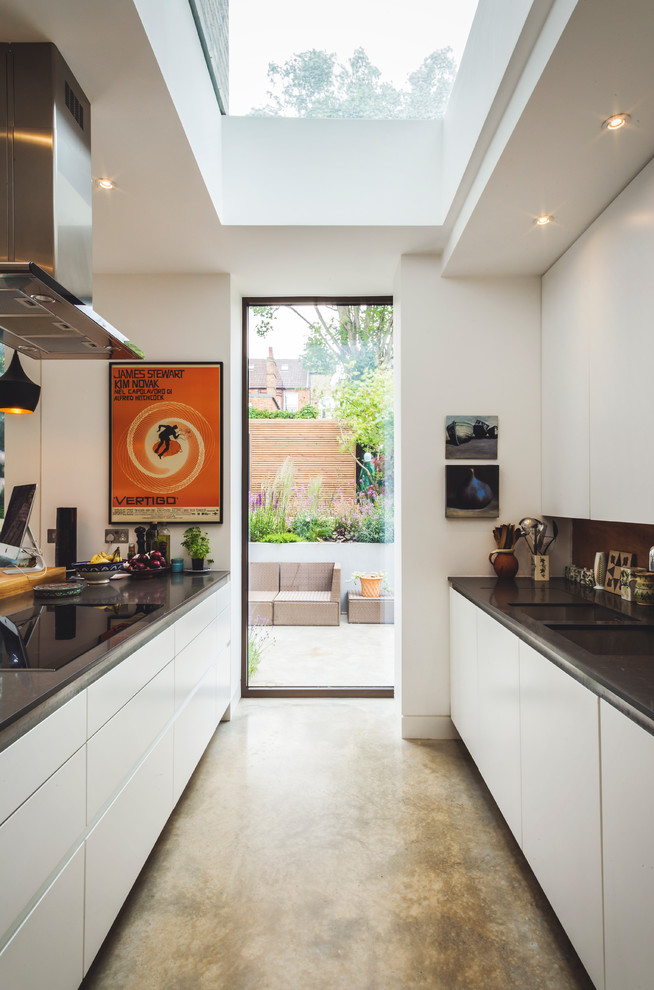 Cette photo montre une cuisine parallèle tendance de taille moyenne avec un placard à porte plane, des portes de placard blanches, sol en béton ciré, plan de travail noir et plafond verrière.