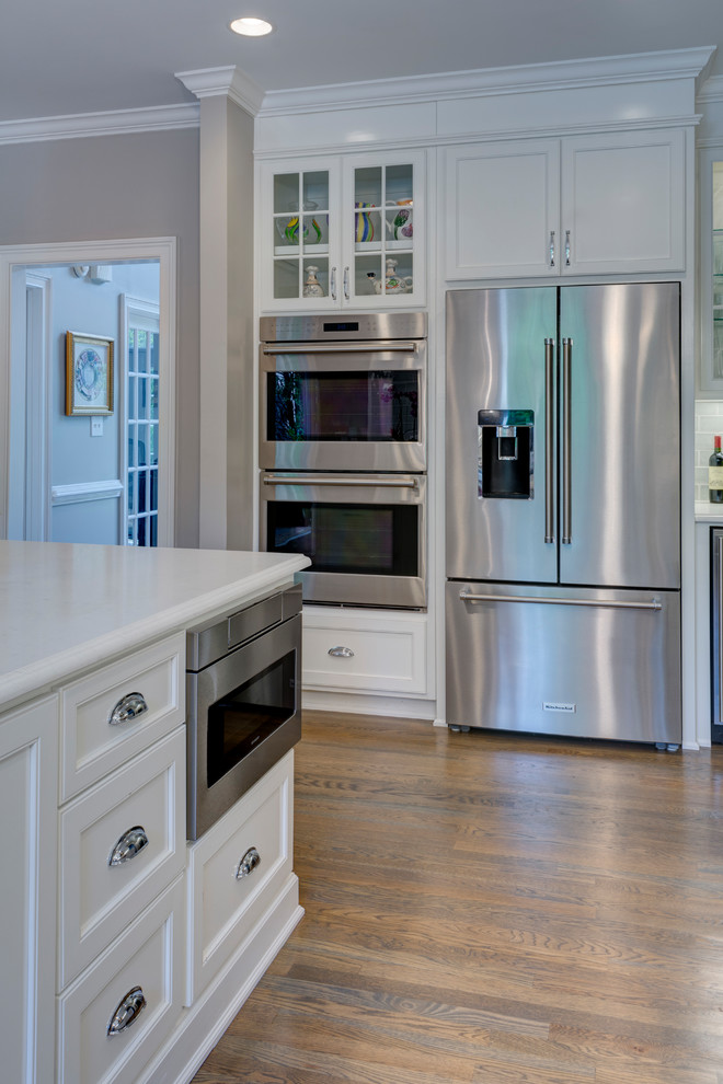 Example of a large transitional u-shaped medium tone wood floor and gray floor eat-in kitchen design in Atlanta with a farmhouse sink, recessed-panel cabinets, white cabinets, quartz countertops, gray backsplash, porcelain backsplash, stainless steel appliances and two islands