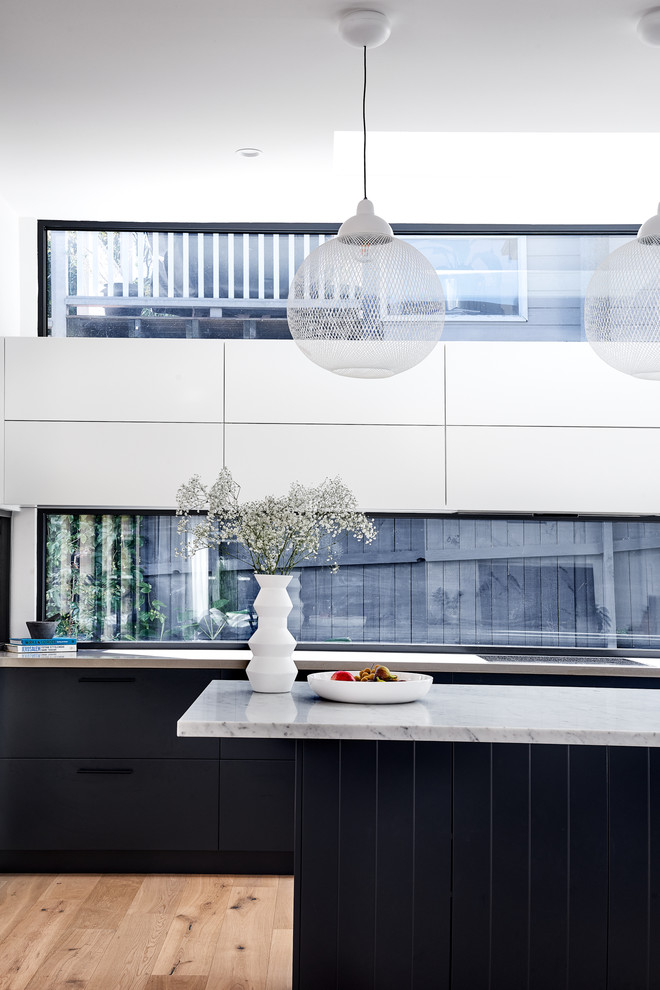 Modern inredning av ett mellanstort blå linjärt blått kök med öppen planlösning, med en undermonterad diskho, blå skåp, marmorbänkskiva, fönster som stänkskydd, ljust trägolv och en köksö
