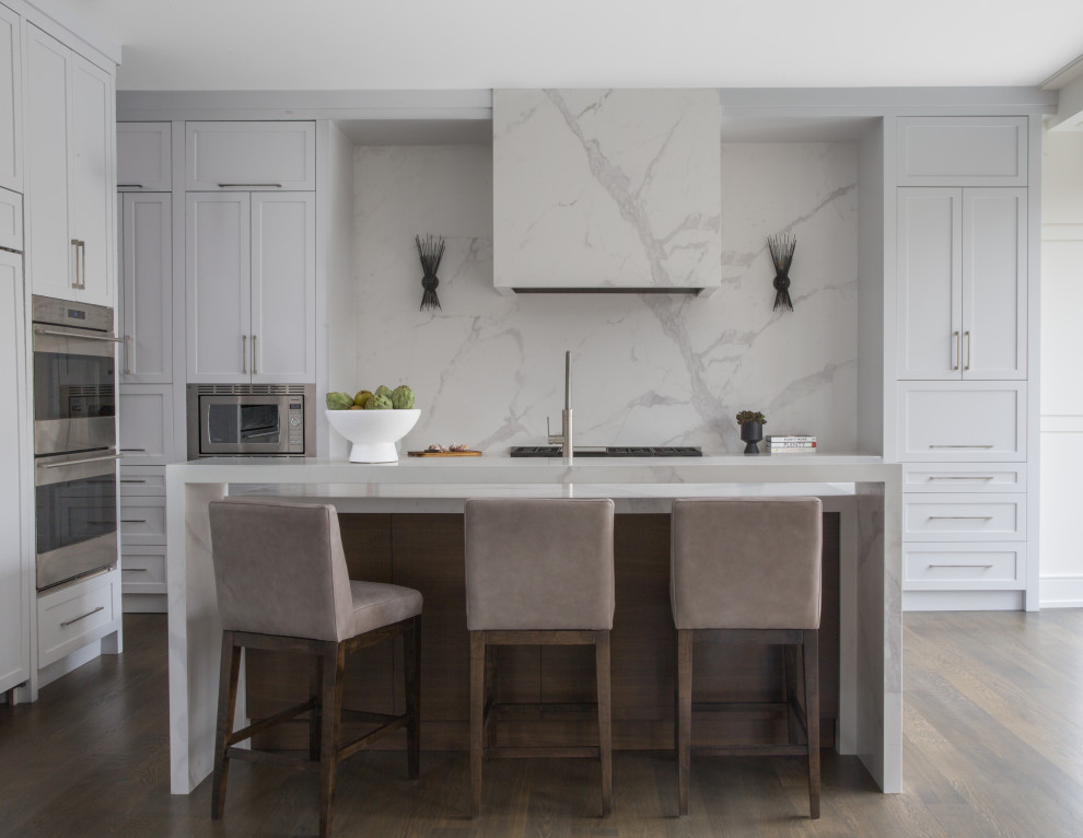 Inredning av ett modernt vit vitt l-kök, med skåp i shakerstil, vita skåp, vitt stänkskydd, stänkskydd i sten, rostfria vitvaror, mörkt trägolv, en köksö och brunt golv