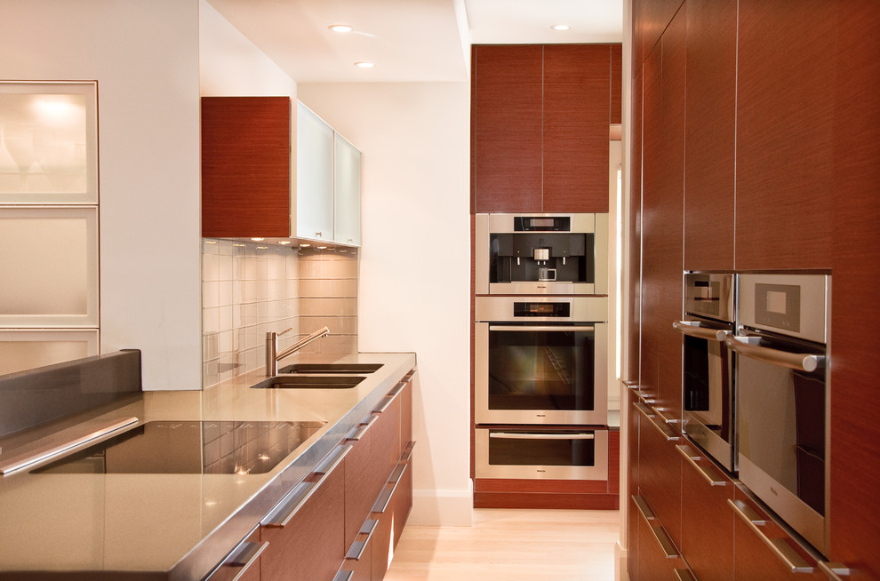 Ejemplo de cocina estrecha contemporánea con electrodomésticos de acero inoxidable, armarios con paneles lisos, puertas de armario de madera en tonos medios y salpicadero de azulejos de vidrio