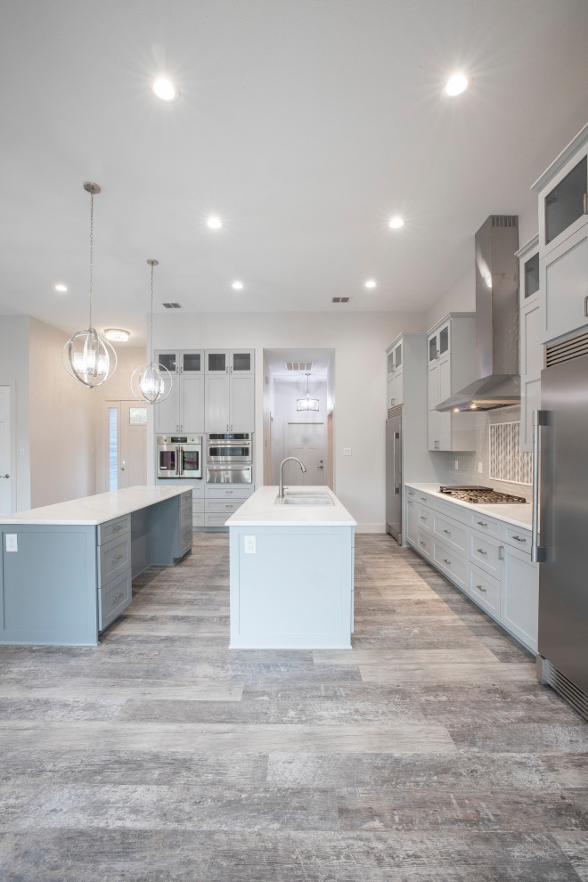 Modern inredning av ett stort vit vitt kök, med grå skåp, granitbänkskiva, grått stänkskydd, stänkskydd i glaskakel, flera köksöar, rostfria vitvaror, en trippel diskho, skåp i shakerstil, vinylgolv och grått golv