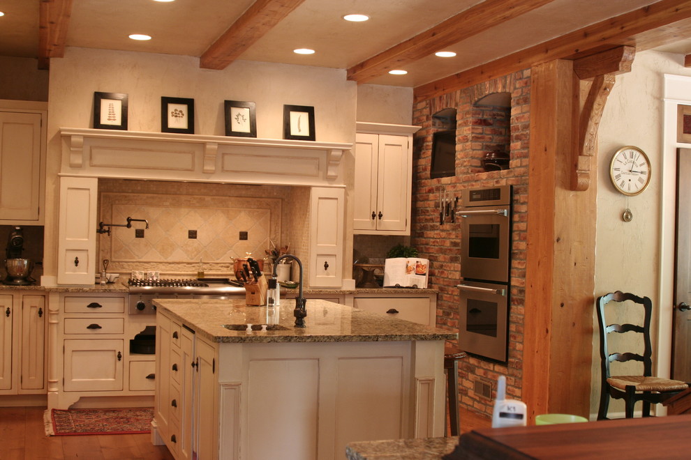 Foto på ett mellanstort lantligt brun kök, med vita skåp, beige stänkskydd, rostfria vitvaror, mellanmörkt trägolv, en undermonterad diskho, skåp i shakerstil, granitbänkskiva, stänkskydd i travertin, en köksö och brunt golv