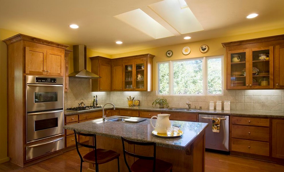 Cette photo montre une cuisine américaine tendance en L et bois brun avec un évier posé, un placard à porte vitrée, un plan de travail en granite, une crédence beige, une crédence en terre cuite et un électroménager en acier inoxydable.
